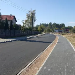 Nowa asfaltowa droga z szarym chodnikiem z kostki brukowej, oddzielona pasem żwiru, w krajobrazie podmiejskim z domami i drzewami w słoneczny dzień.