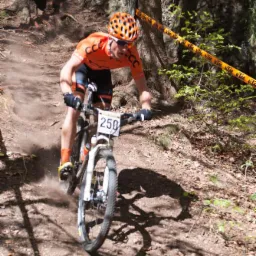 Kolarz górski w pomarańczowej koszulce i kasku, z numerem 250 na rowerze, pokonuje stromy, leśny zjazd podczas wyścigu, otoczony taśmą wyznaczającą trasę.