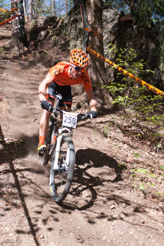 Kolarz górski w pomarańczowej koszulce i kasku, z numerem 250 na rowerze, pokonuje stromy, leśny zjazd podczas wyścigu, otoczony taśmą wyznaczającą trasę.