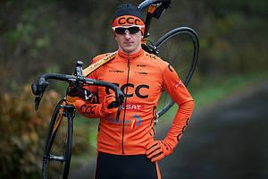 Mężczyzna w pomarańczowym stroju kolarskim z logo CCC, trzymający rower na ramieniu, okulary przeciwsłoneczne, czapka z daszkiem, leśne tło.