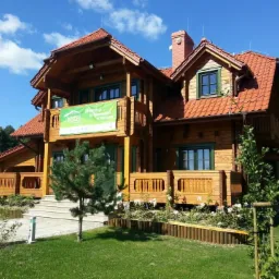 Gotowy, dwukondygnacyjny dom z bali z czerwonym dachem, zielonymi ramami okiennymi i balkonem, otoczony zielenią i ogrodem z młodymi drzewkami.