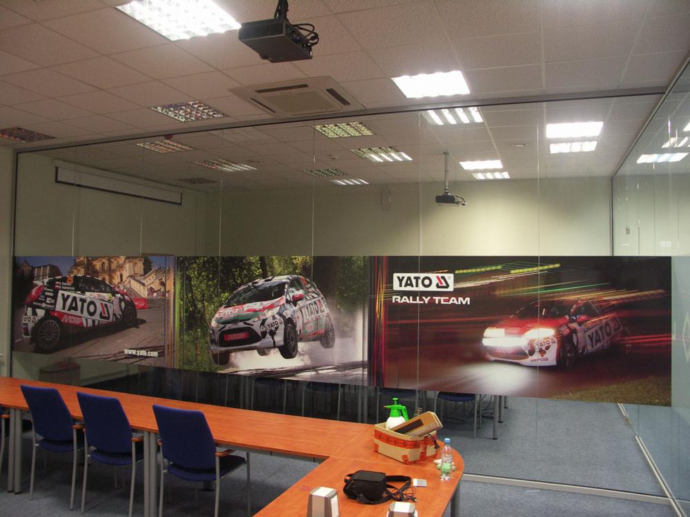 Wnętrze biura z powieszonymi banerami reklamowymi YATO Rally Team przedstawiającymi samochody rajdowe w akcji, widoczne stoły konferencyjne i krzesła.