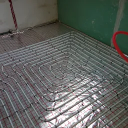 Białe rury ogrzewania podłogowego ułożone na folii izolacyjnej z czerwoną siatką, przymocowane czarnymi uchwytami, z fragmentem czerwonej rury przy ścianie w surowym wnętrzu.