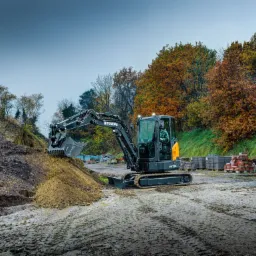 Czarna maszyna budowlana Hyundai HX35Az, prawdopodobnie minikoparka, nabiera ziemię z pryzmy na błotnistym terenie w Liszkach, w tle drzewa z jesiennymi liśćmi i palety z kostką brukową.
