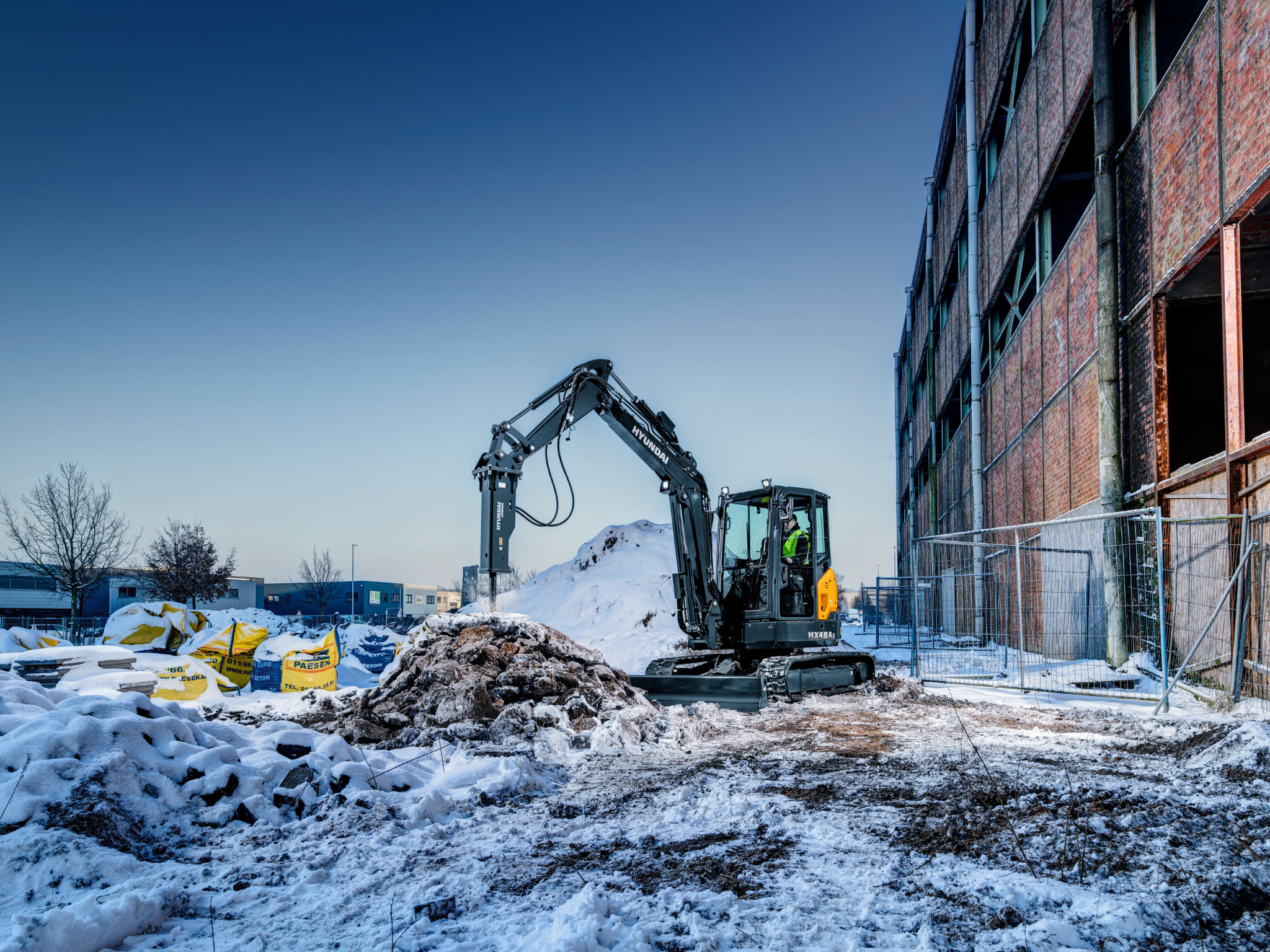 Czarna minikoparka Hyundai HX48Az z młotem hydraulicznym rozbija zamarzniętą ziemię na placu budowy w zimowy dzień. W tle sterta worków z materiałami budowlanymi i budynek w trakcie renowacji.