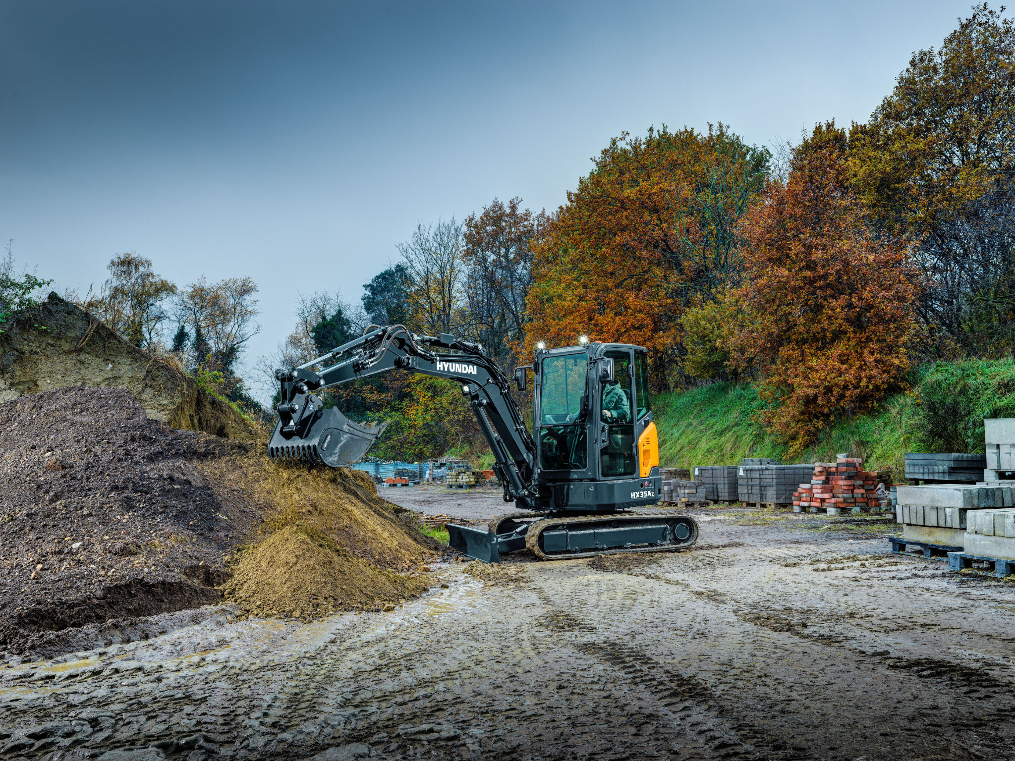 Czarna maszyna budowlana Hyundai HX35Az, prawdopodobnie minikoparka, nabiera ziemię z pryzmy na błotnistym terenie w Liszkach, w tle drzewa z jesiennymi liśćmi i palety z kostką brukową.