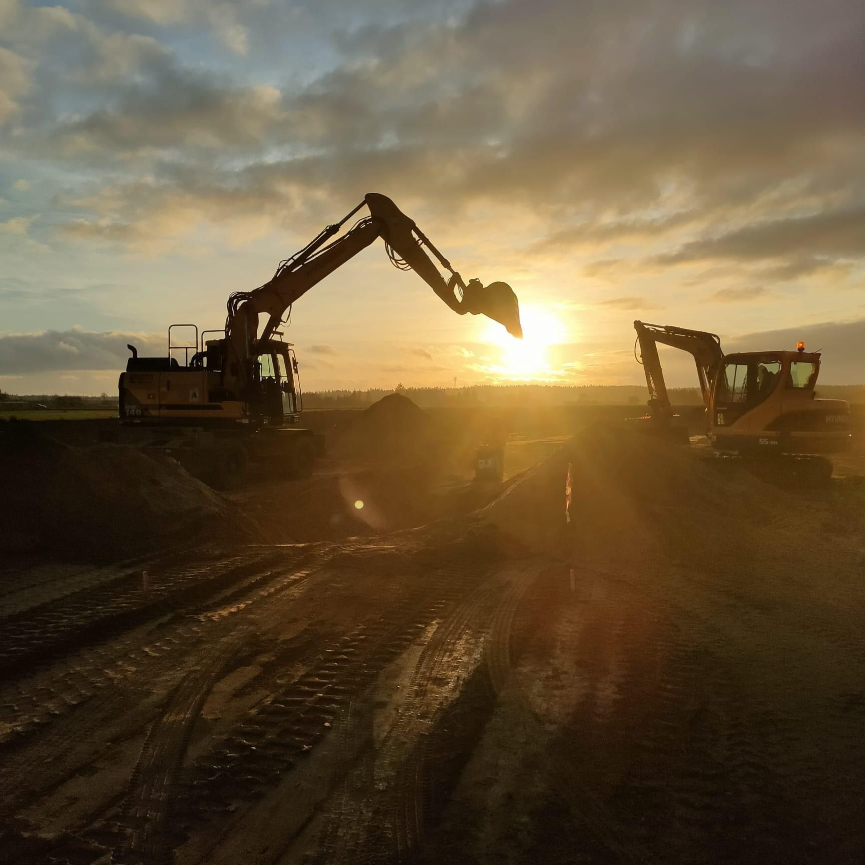 Dwie koparki w silnych promieniach zachodzącego słońca, tworzące sylwetki na tle nieba z chmurami, ślady opon na ziemi.