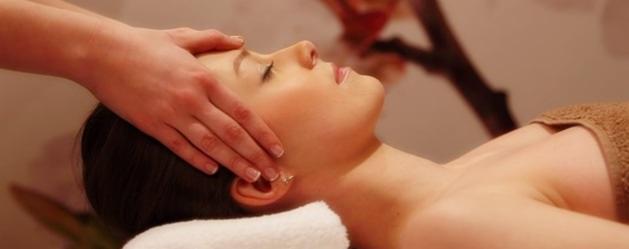 Kobieta leżąca na białym ręczniku podczas masażu głowy, dłonie terapeuty delikatnie uciskają skronie, w tle rozmyty motyw kwiatowy.