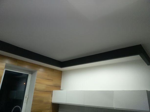 Nowoczesna kuchnia po remoncie: białe szafki wiszące, drewnopodobne płytki na ścianie, czarny dekoracyjny element pod sufitem.