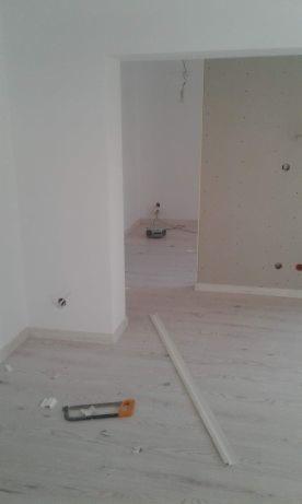 Wnętrze w trakcie remontu: widoczna nowa podłoga imitująca drewno, białe ściany, listwa przypodłogowa i piła ręczna leżące na podłodze, w tle pomieszczenie z płytą kartonowo-gipsową.