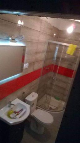 Ukończona łazienka z kabiną prysznicową, umywalką z miską na akcesoria i toaletą, wykończona szarymi płytkami z czerwonym pasem dekoracyjnym.