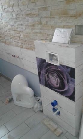 Montaż stelaża podtynkowego WC z dekoracyjną grafiką kwiatową, widoczne przyłącza, płytki imitujące kamień na ścianie, surowy stan wykończenia.