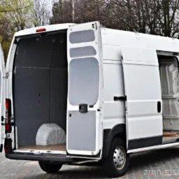 Biały furgon z otwartymi tylnymi i bocznymi drzwiami, ukazujący puste wnętrze przestrzeni ładunkowej, na tle drzew.