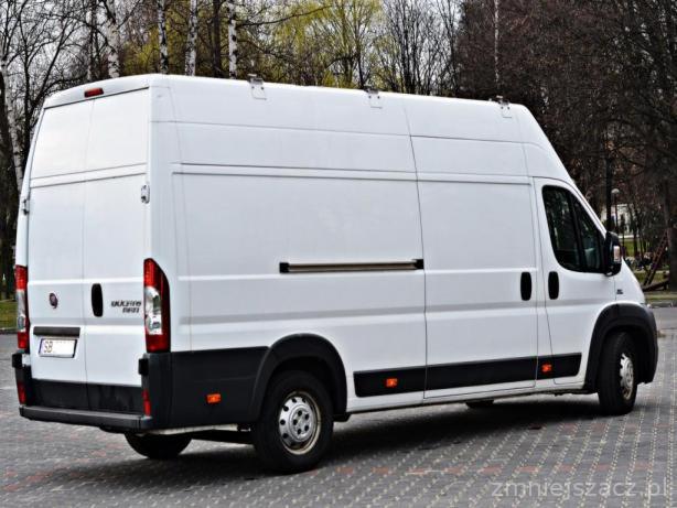 Biały furgon dostawczy Fiat Ducato z przesuwnymi drzwiami bocznymi, zaparkowany na kostce brukowej, widok z tyłu i boku.