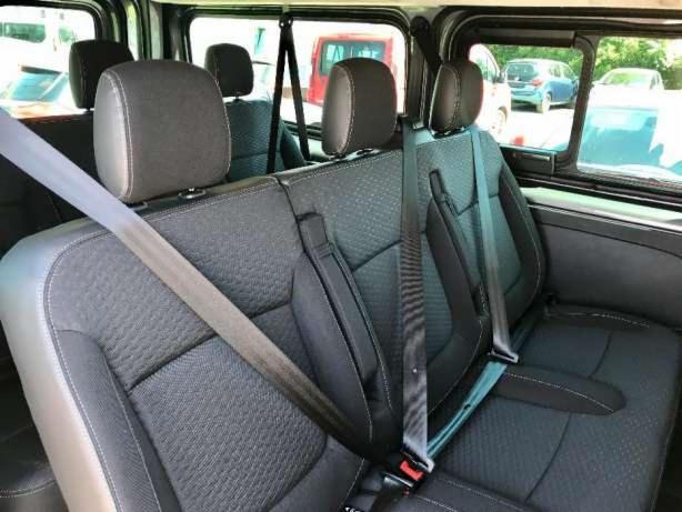 Wnętrze samochodu dostawczego z trzema rzędami foteli z ciemną tapicerką i widocznymi pasami bezpieczeństwa, widok z perspektywy tylnych siedzeń na okna i inne zaparkowane pojazdy na zewnątrz.