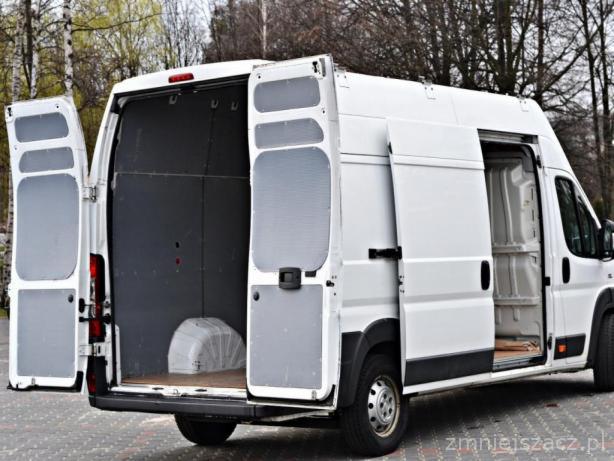 Biały furgon z otwartymi tylnymi i bocznymi drzwiami, ukazujący puste wnętrze przestrzeni ładunkowej, na tle drzew.