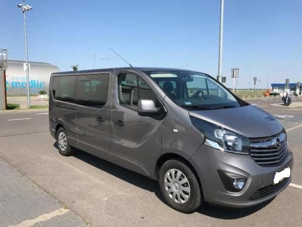 Szary van Opel Vivaro z anteną dachową na parkingu, widoczny budynek lotniska w tle.