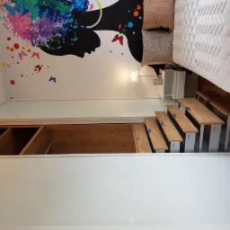 Nowoczesna sypialnia z zabudowaną szafą przesuwną z białym frontem, antresolą z drewnianymi stopniami i kolorową grafiką na ścianie przedstawiającą profil kobiety.