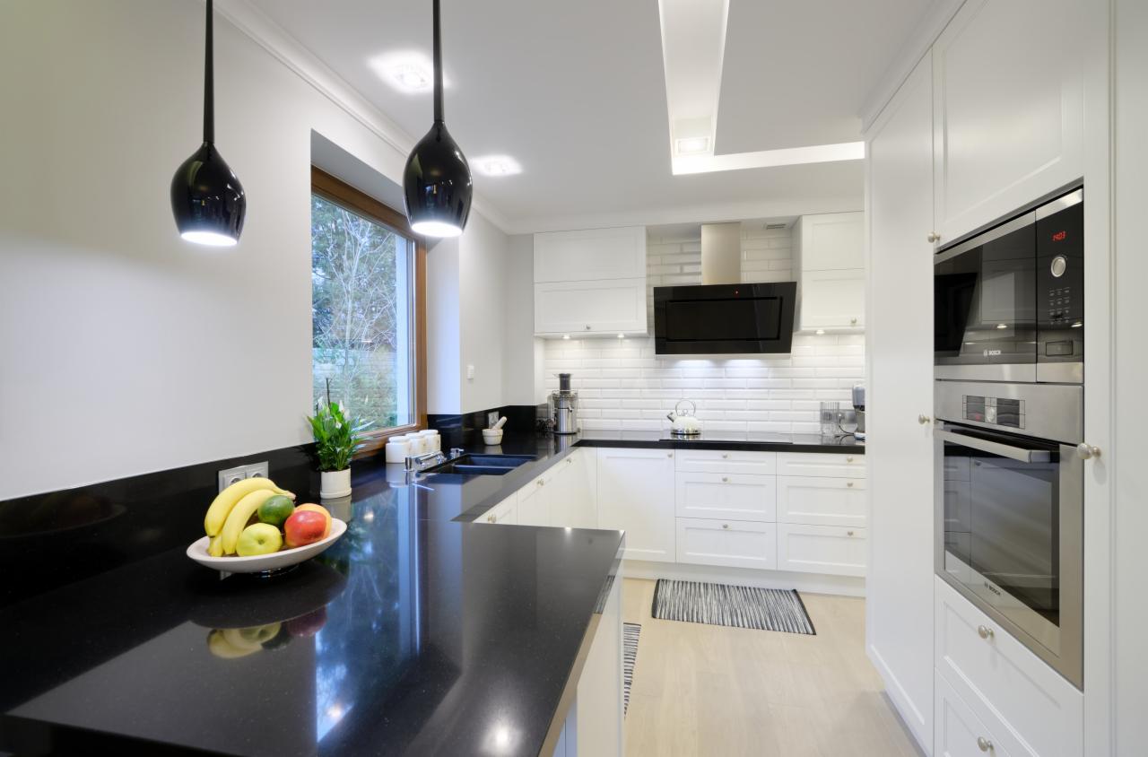 Biała kuchnia z czarnym blatem i owocami w misie. Nad blatem wiszą czarne lampy, a w tle widać okno. Nowoczesny design, oświetlenie LED, czysty kadr.