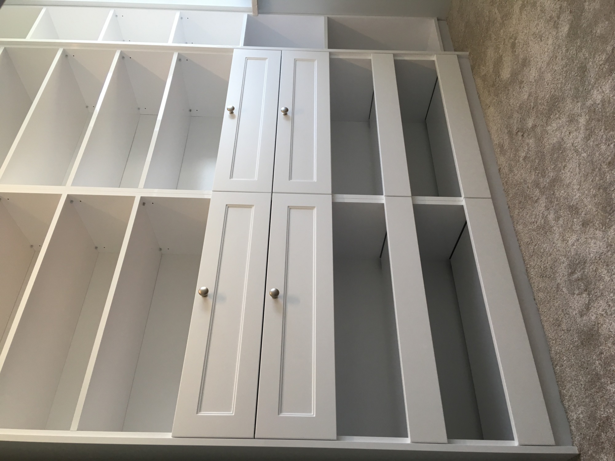 Biała, częściowo zabudowana szafa z otwartymi półkami i czterema drzwiczkami z metalowymi gałkami, widoczna na tle beżowej wykładziny.