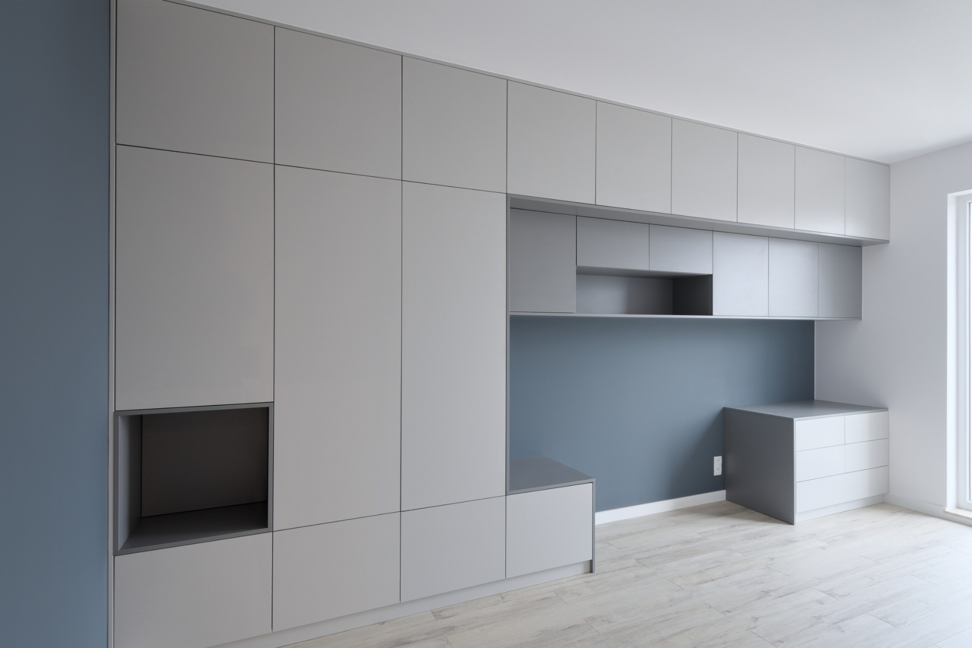 Jasnoszara zabudowa meblowa z szafkami górnymi i otwartą wnęką, uzupełniona biurkiem z szufladami przy ścianie w odcieniu błękitu.