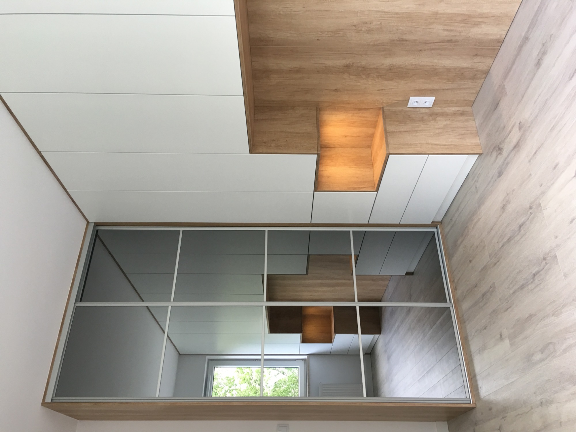 Nowoczesna szafa wnękowa z przesuwnymi drzwiami lustrzanymi, odbijająca fragment sufitu z drewnianymi elementami i oświetleniem punktowym, widok z dołu.