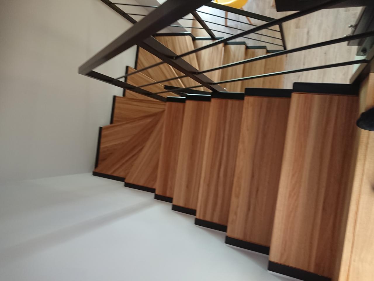 Spiralne schody z drewnianymi stopniami i czarną metalową balustradą, widok z góry, z jasnym oświetleniem podkreślającym teksturę drewna.