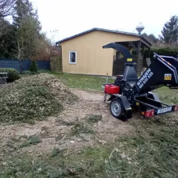 Zrębkowanie gałęzi na zielonym podwórku. Czarna maszyna do zrębkowania z numerem rejestracyjnym ZKA 92EE stoi obok sterty zrębków. W tle żółty budynek i drzewa.