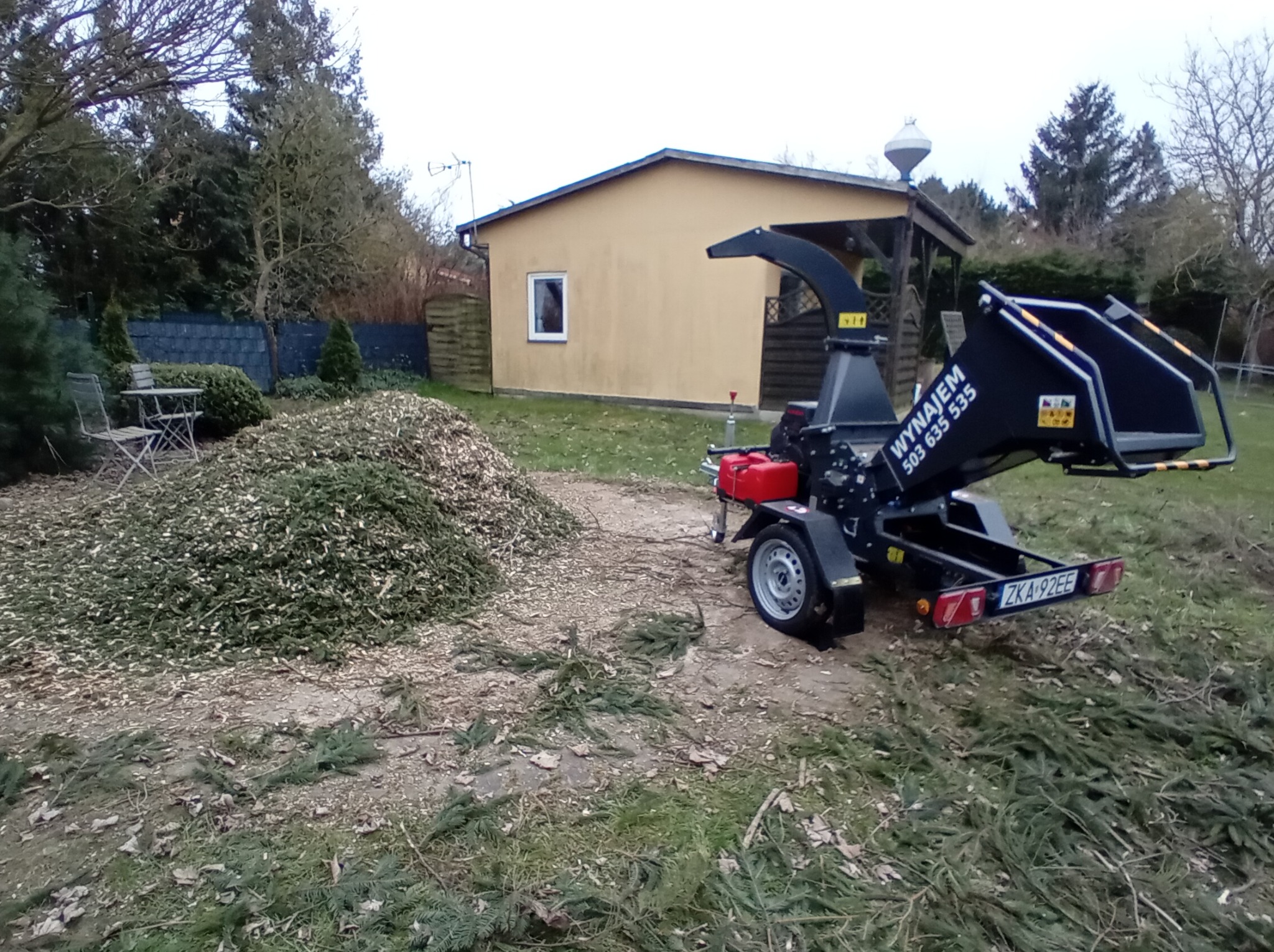 Zrębkowanie gałęzi na zielonym podwórku. Czarna maszyna do zrębkowania z numerem rejestracyjnym ZKA 92EE stoi obok sterty zrębków. W tle żółty budynek i drzewa.