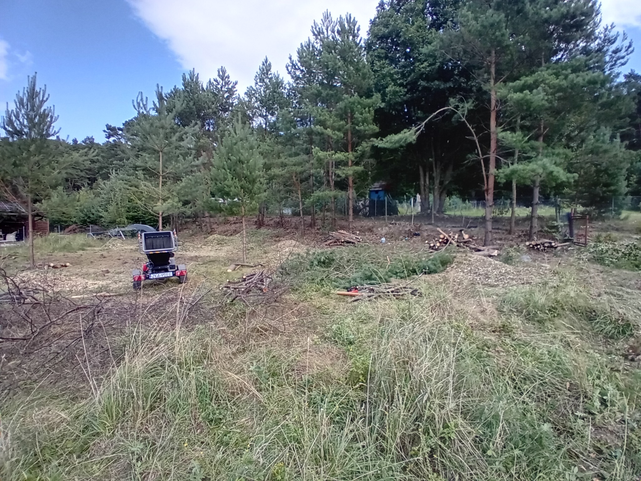 Teren po wycince drzew z widoczną rębarką do gałęzi i stertami pociętych gałęzi. W tle drzewa iglaste i fragment budynku.