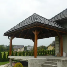 Zadaszona konstrukcja z drewnianymi słupami i betonową podstawą, przylegająca do domu z ciemną dachówką, widok na ogród i sąsiednie budynki w tle.