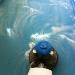 Maszyna do czyszczenia podłóg w trakcie pracy na niebieskiej, błyszczącej powierzchni, pozostawiająca ślady piany i wody.