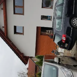 Jasna elewacja domu z drewnianymi elementami wykończeniowymi, widoczne zaparkowane samochody i drabina przy garażu.