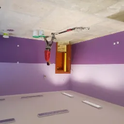 Mężczyzna w czerwonej koszuli maluje ścianę na fioletowo za pomocą szlifierki do gipsu; widoczne puszki z farbą i niedokończony sufit.