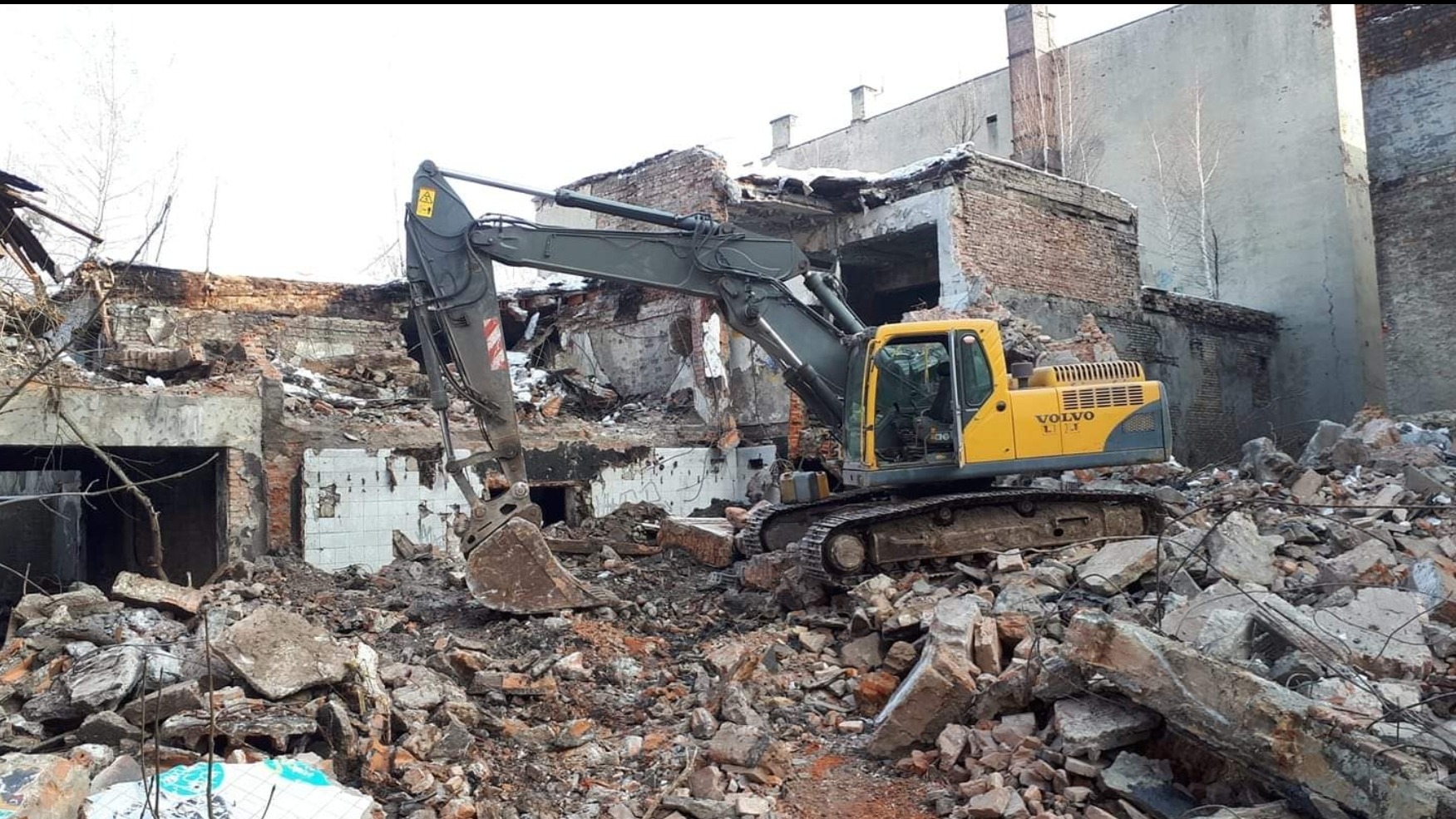 Żółta koparka Volvo na gąsienicach stoi na stosie gruzu po rozbiórce budynku, w tle fragmenty ceglanej ściany i białe płytki.