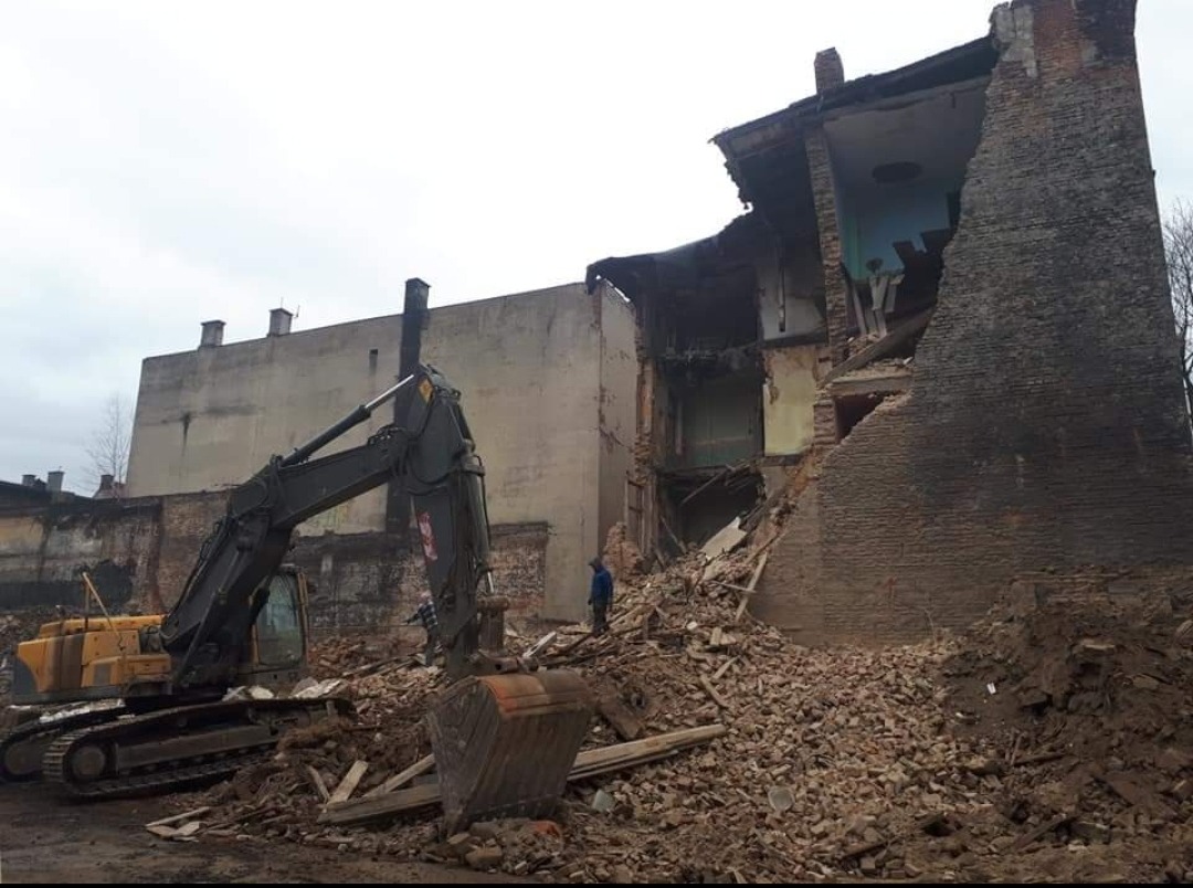Żółta koparka gąsienicowa na tle częściowo zburzonego, ceglanego budynku z odsłoniętymi fragmentami wnętrz i stertą gruzu.