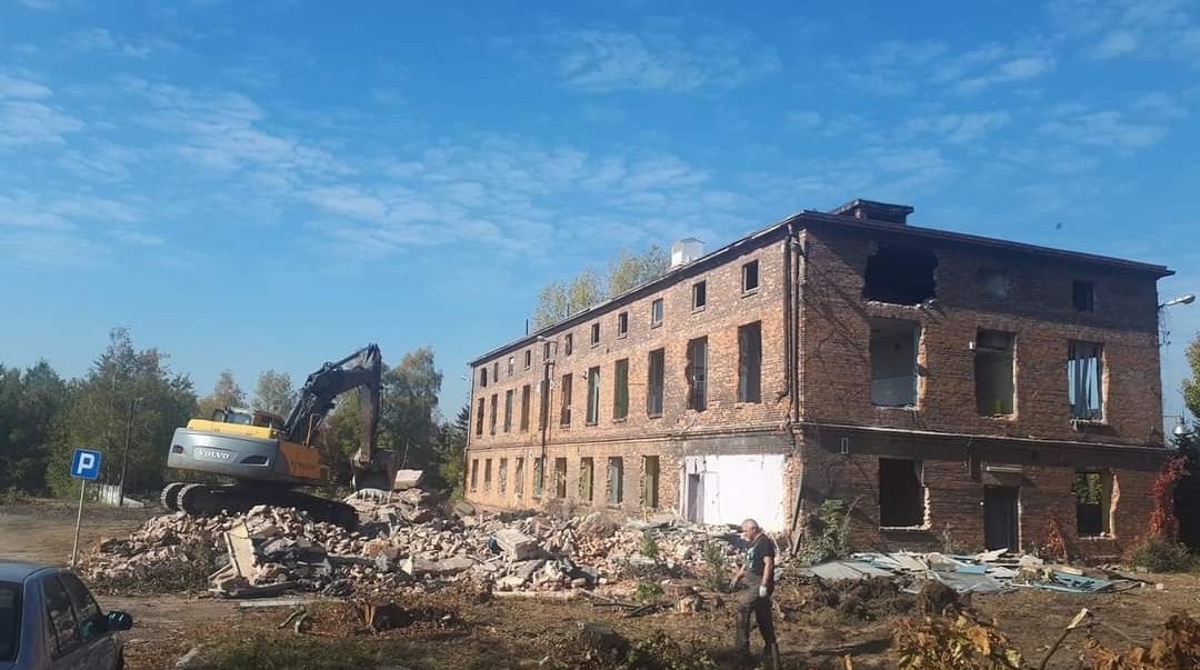 Żółto-czarna koparka Volvo na tle częściowo zburzonego, ceglanego budynku z powybijanymi oknami i gruzem, widoczny znak drogowy P i fragment auta. W tle drzewa i błękitne niebo.