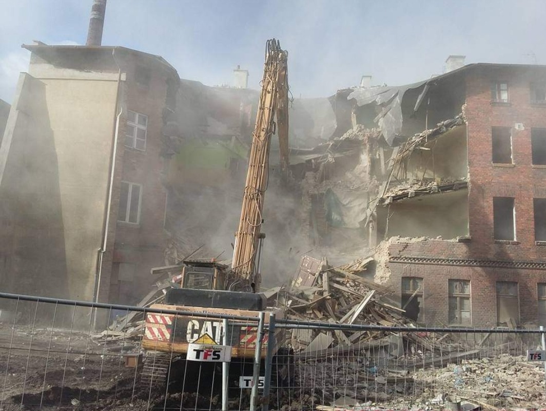Ciężki sprzęt CAT z długim ramieniem burzy ceglany budynek, unoszący się pył i gruz, widoczne zabezpieczenia placu budowy.