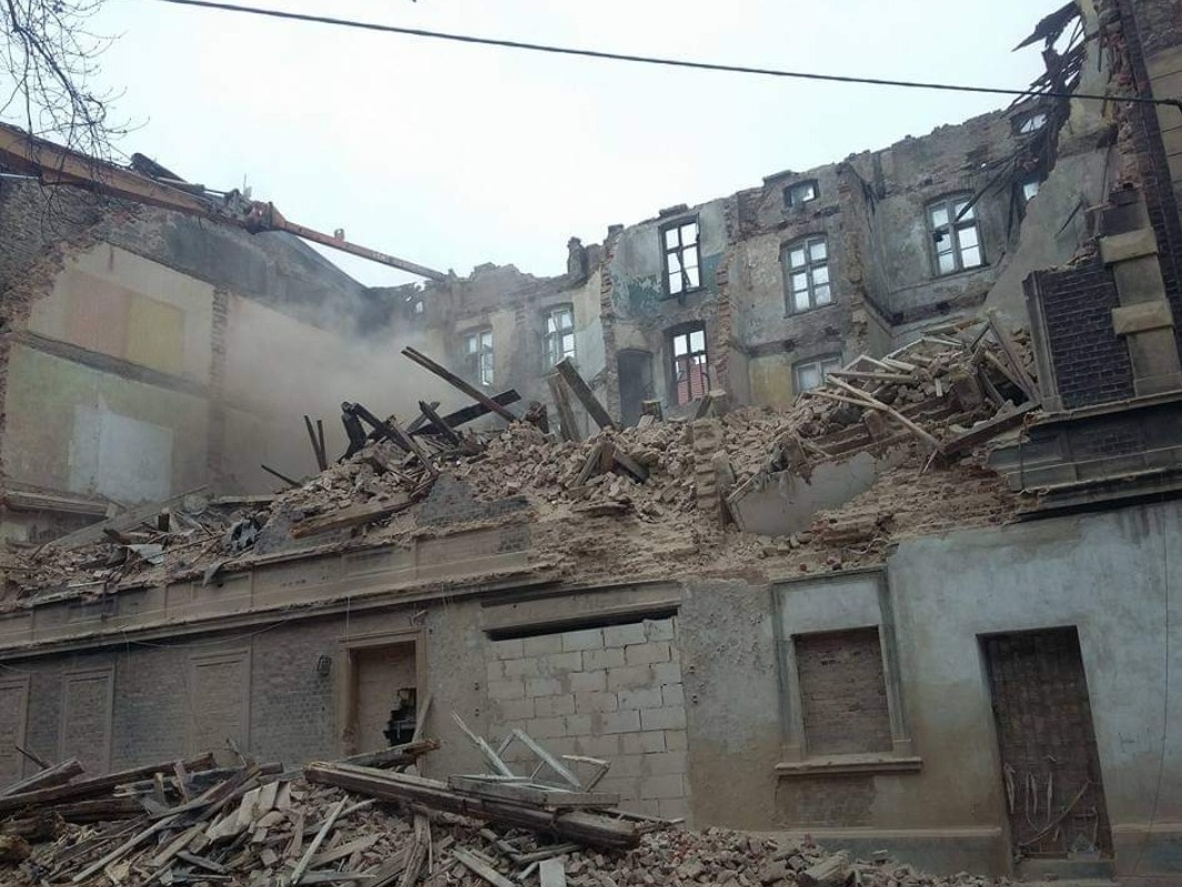 Częściowo zburzony budynek z gruzem i elementami konstrukcyjnymi, widoczny żuraw budowlany w tle, unoszący się pył.