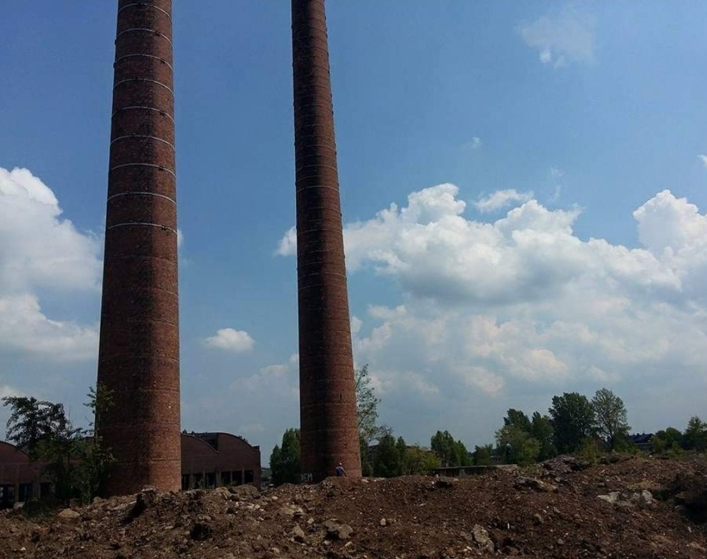 Dwa wysokie, ceglane kominy na tle błękitnego nieba z obłokami, teren po wyburzeniu z widocznymi resztkami gruzu i fragmentami budynku w tle.