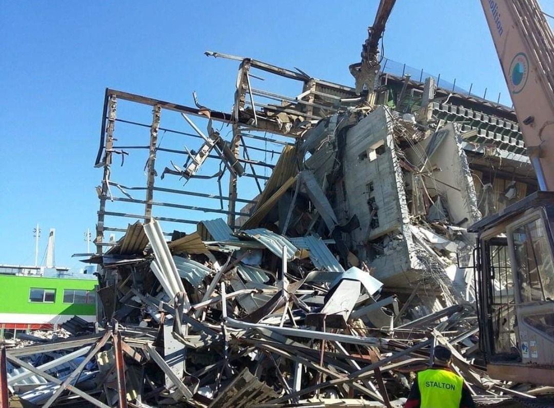 Częściowo zburzony budynek z elementami stalowej konstrukcji, gruzem i fragmentami betonu, na pierwszym planie pracownik w żółtej kamizelce odblaskowej, w tle zielony budynek i błękitne niebo.