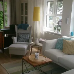 Jasny salon z elementami skandynawskimi: biała sofa z poduszkami w geometryczne wzory, fotel uszak z podnóżkiem, dywan w pasy i stolik kawowy z drewnianym blatem na metalowym stelażu.