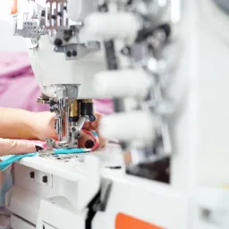 Zbliżenie na proces szycia na overlocku; dłonie przesuwają błękitny materiał pod stopką maszyny, widoczne metalowe elementy maszyny i fragmenty kolorowej tkaniny w tle.