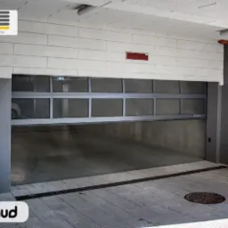 Częściowo otwarta segmentowa brama do hali garażowej z aluminiową ramą i przeszklonymi panelami, widoczna sygnalizacja świetlna nad bramą, posadzka z szarej kostki brukowej z odpływem liniowym...