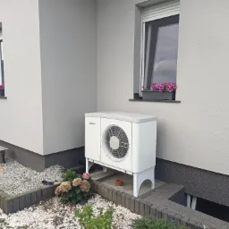 Biała pompa ciepła marki Stiebel Eltron zamontowana przy ścianie domu z dwoma oknami, w otoczeniu kamieni i roślin.