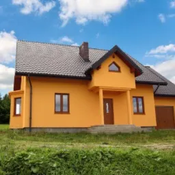 Ocieplony dom jednorodzinny z pomarańczową elewacją, brązową stolarką okienną i grafitową dachówką na tle błękitnego nieba z chmurami.