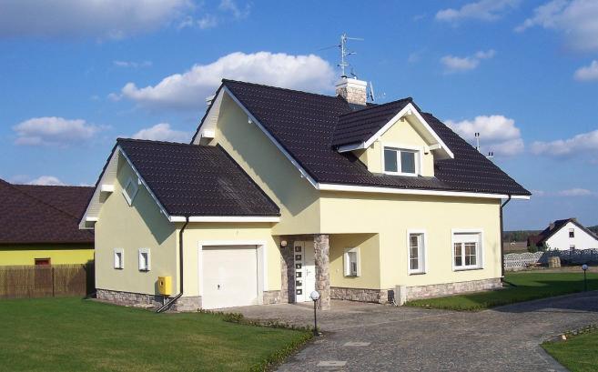 Żółty dom jednorodzinny z ciemnym dachem i garażem, wykończony kamieniem elewacyjnym u podstawy, na tle błękitnego nieba z chmurami.