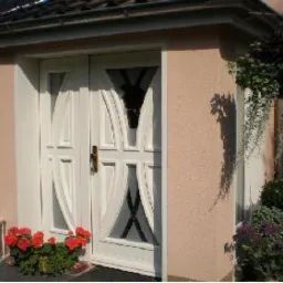 Białe, podwójne drzwi wejściowe z dekoracyjnymi szybami i zasłonkami, umieszczone w osłoniętym wejściu do domu, otoczone roślinami i latarenką.