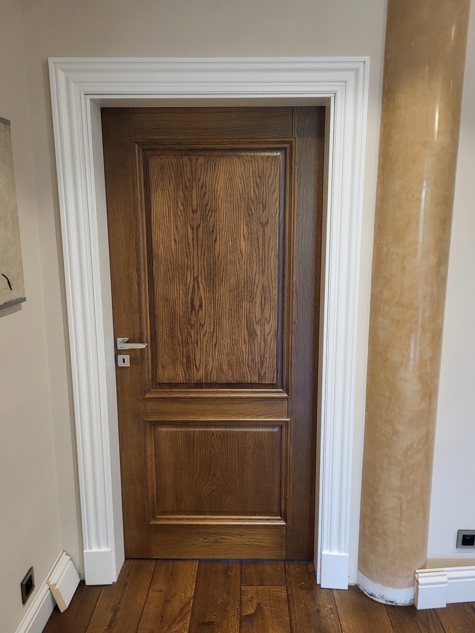 Klasyczne drewniane drzwi wewnątrz pomieszczenia, z dekoracyjną białą ramą i kolumną w tle, widoczny fragment podłogi z paneli.