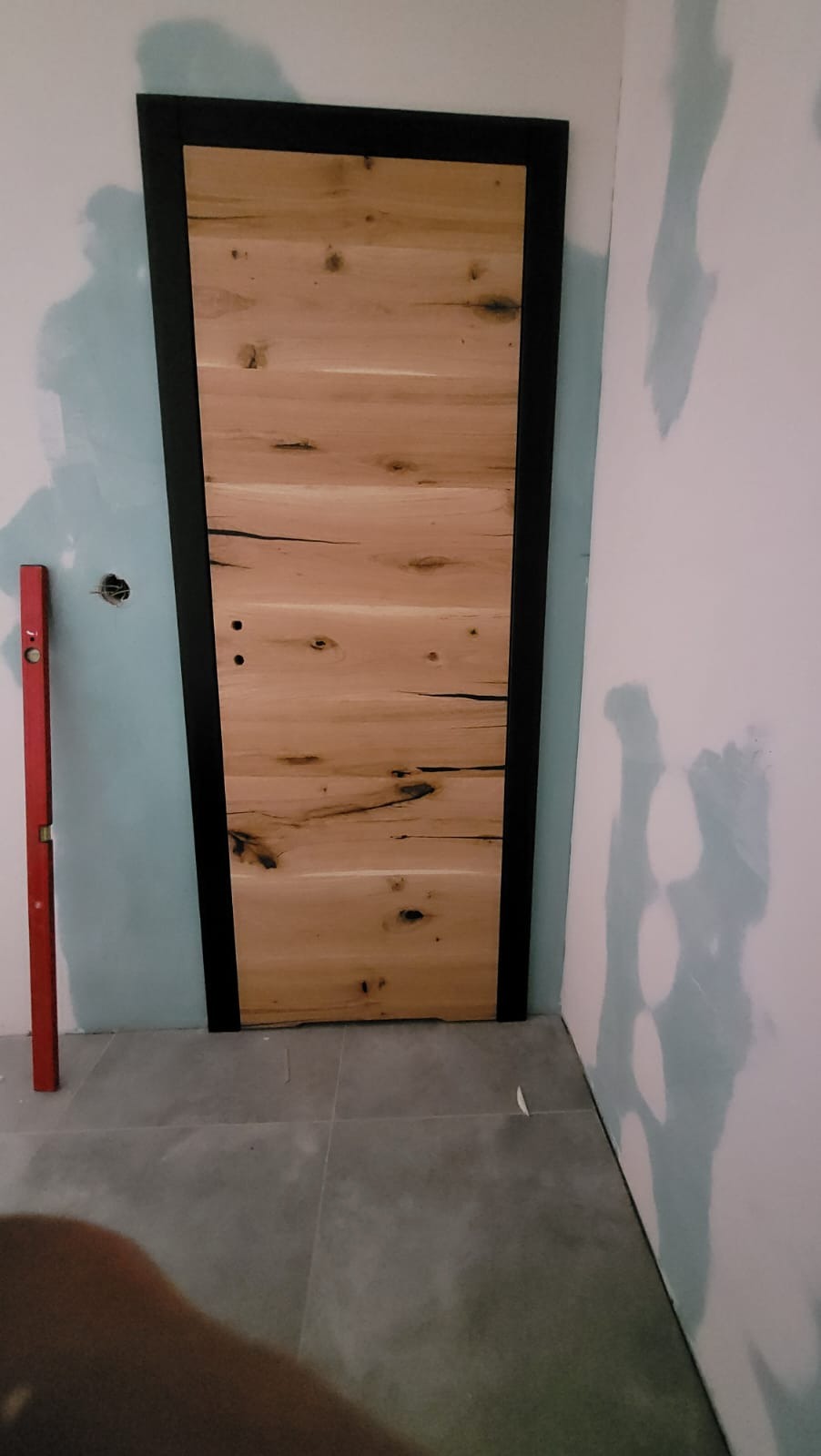 Montaż drzwi z naturalnego drewna z czarną ramą, widoczny poziomica na ścianie, surowy stan wykończenia wnętrza.